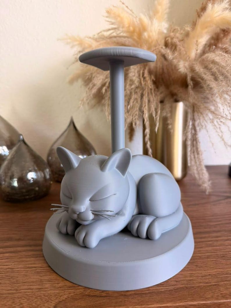 May include: A light gray paper towel holder featuring a sculpted sleeping cat on a circular base. The cat has closed eyes and visible whiskers, with a vertical pole extending from its back to support a horizontal top disc for holding rolls.