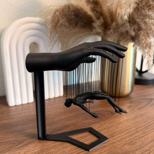May include: A black metal sculpture featuring a hand and a figure suspended by strings. The hand is positioned above the figure, which appears to be in a diving pose. The sculpture is on a wooden surface.