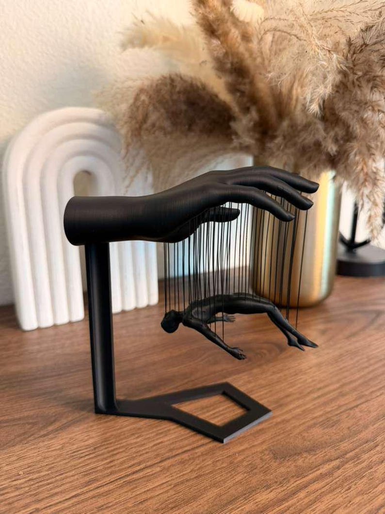 May include: A black metal sculpture featuring a hand and a figure suspended by strings. The hand is positioned above the figure, which appears to be in a diving pose. The sculpture is on a wooden surface.