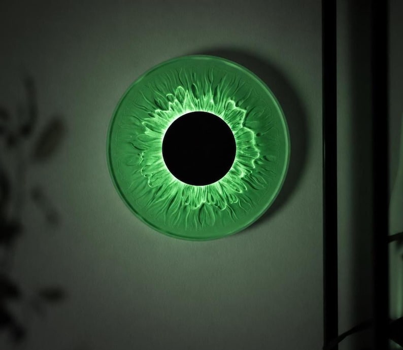 May include: A circular wall light fixture featuring a solid black center disc surrounded by a translucent, textured green ring that radiates light outward, creating a pattern reminiscent of a human iris or a stylized sunflower bloom.