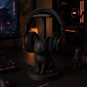 May include: A matte black headphone stand featuring a sculpted cat base with closed eyes and whiskers. The stand includes a vertical post and a top horizontal bar designed to hold a pair of over-ear headphones securely in place.
