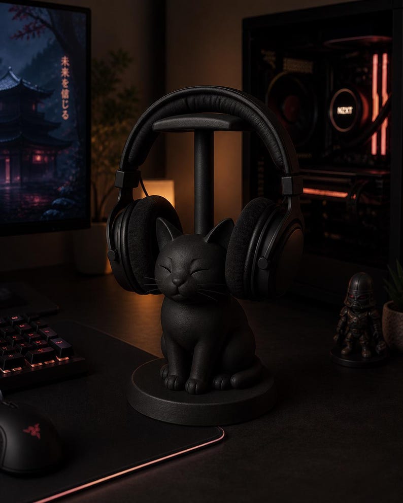 May include: A matte black headphone stand featuring a sculpted cat base with closed eyes and whiskers. The stand includes a vertical post and a top horizontal bar designed to hold a pair of over-ear headphones securely in place.