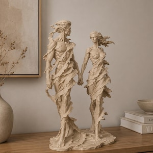 May include: A beige sculpture depicting two figures standing side by side and holding hands. The figures are draped in flowing, tattered fabric that appears to be caught in a breeze, revealing their muscular forms. The piece has a matte finish.