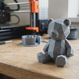 May include: A gray, geometric-style teddy bear figurine sits on a wooden surface. The bear is made of many flat, triangular surfaces. A 3D printer and other tools are in the background.