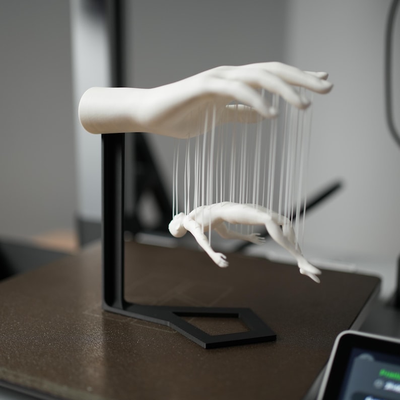 May include: A white sculpture of a hand holding a marionette figure suspended by strings. The hand and figure are white, with a black metal stand. The figure is in a diving pose.
