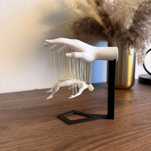 May include: A white hand sculpture with a black base, holding a small figure suspended by strings. The sculpture is on a wooden surface, with a vase of dried flowers in the background.