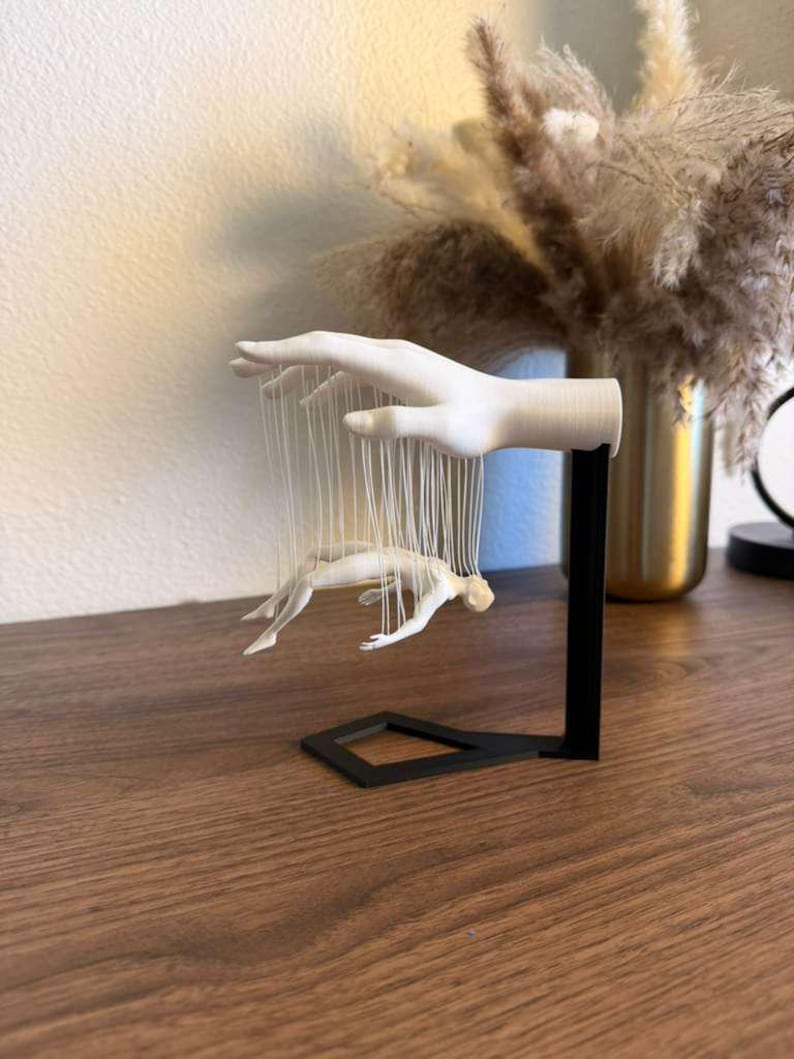 May include: A white hand sculpture with a black base, holding a small figure suspended by strings. The sculpture is on a wooden surface, with a vase of dried flowers in the background.