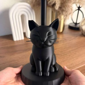 May include: A matte black paper towel holder featuring a sculpted cat sitting on a circular base. The cat has closed eyes and thin, dark whiskers. A vertical pole extends from the cat's back, topped with a flat, square-shaped support piece.