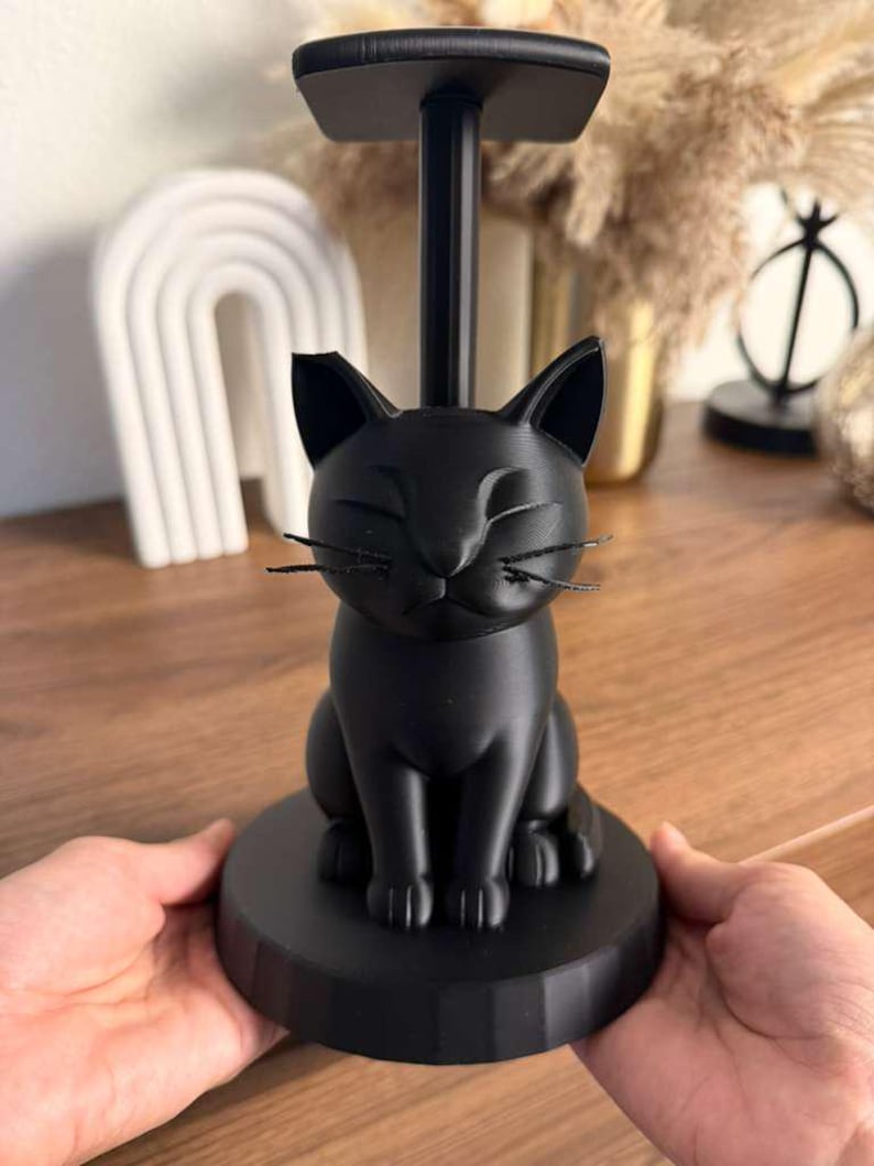 May include: A matte black paper towel holder featuring a sculpted cat sitting on a circular base. The cat has closed eyes and thin, dark whiskers. A vertical pole extends from the cat's back, topped with a flat, square-shaped support piece.