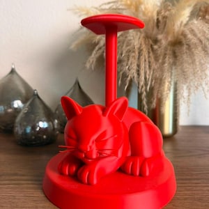 May include: A bright red plastic stand featuring a sculpted sleeping cat resting on a circular base. A vertical post extends upward from the cat's back, topped with a flat, circular platform designed to hold items like headphones or jewelry.