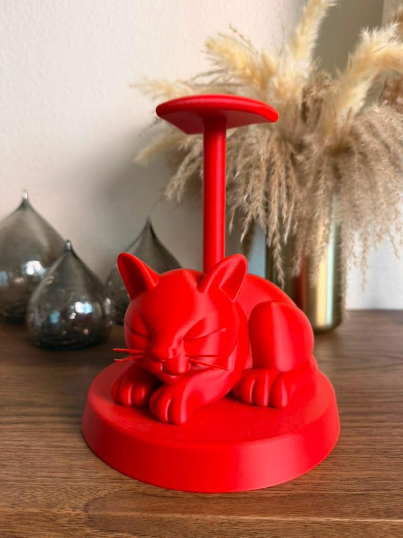 May include: A bright red plastic stand featuring a sculpted sleeping cat resting on a circular base. A vertical post extends upward from the cat's back, topped with a flat, circular platform designed to hold items like headphones or jewelry.