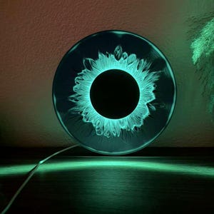 May include: A circular wall lamp featuring a dark, solid black center surrounded by a glowing, vibrant teal ring that mimics the appearance of an iris. The light emits a soft, radiant green illumination that casts a glow onto the surface below.