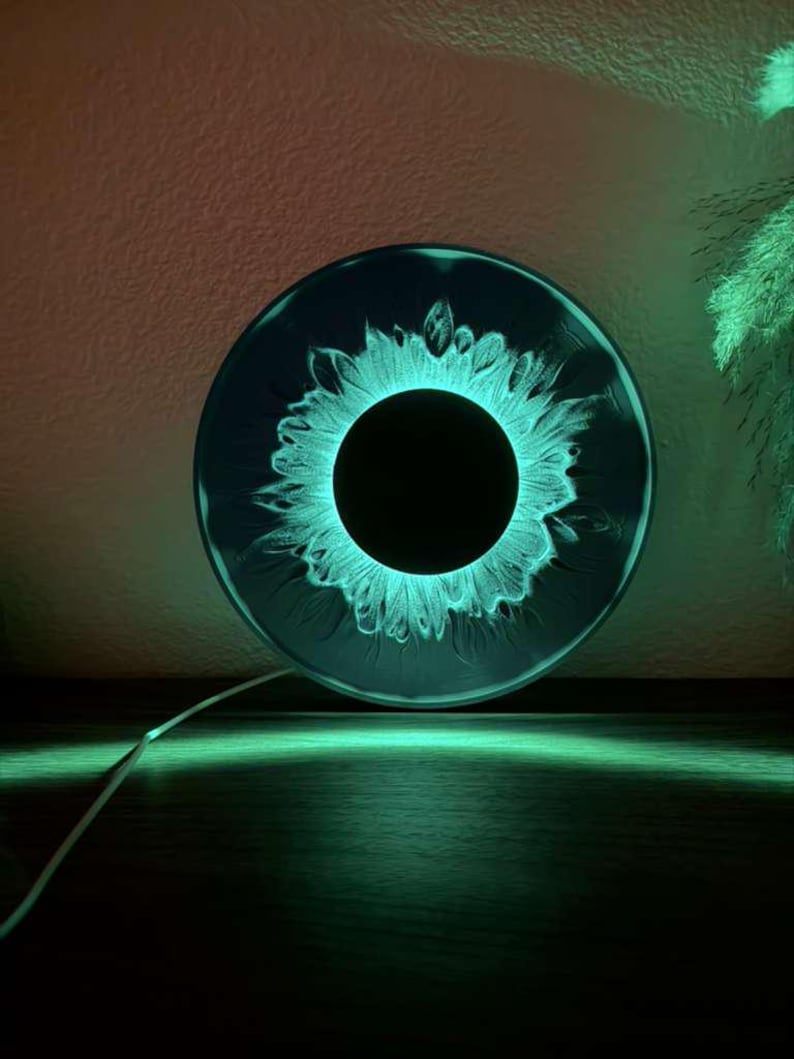 May include: A circular wall lamp featuring a dark, solid black center surrounded by a glowing, vibrant teal ring that mimics the appearance of an iris. The light emits a soft, radiant green illumination that casts a glow onto the surface below.