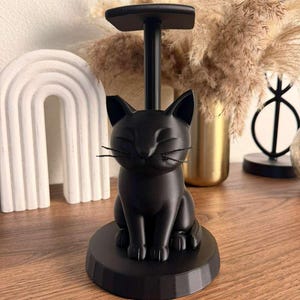 May include: A matte black cat-shaped paper towel holder featuring a sculpted feline figure sitting on a circular base with a vertical rod extending upward to a flat top piece. The cat has pointed ears, closed eyes, and thin black whiskers.