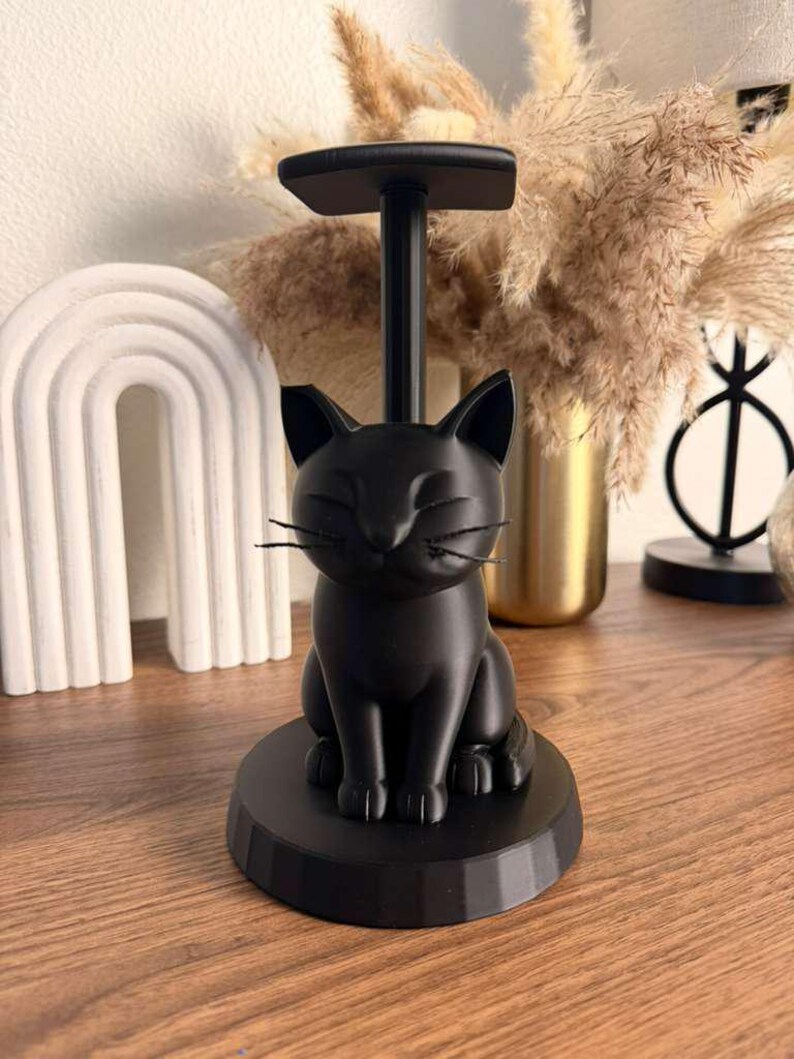 May include: A matte black cat-shaped paper towel holder featuring a sculpted feline figure sitting on a circular base with a vertical rod extending upward to a flat top piece. The cat has pointed ears, closed eyes, and thin black whiskers.