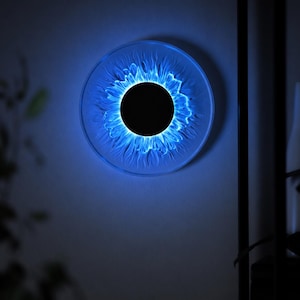 May include: A circular wall light fixture featuring a solid black center disc surrounded by a translucent, textured ring that emits a vibrant blue glow. The light radiates outward from the center, creating a pattern resembling a stylized sun or iris.