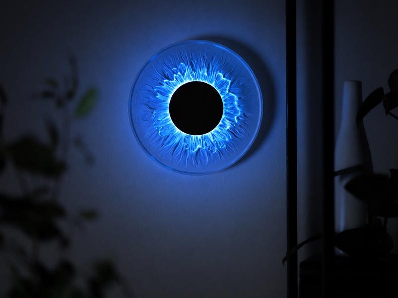 May include: A circular wall light fixture featuring a solid black center disc surrounded by a translucent, textured ring that emits a vibrant blue glow. The light radiates outward from the center, creating a pattern resembling a stylized sun or iris.