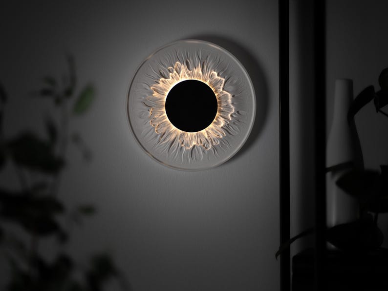 May include: A circular wall light fixture featuring a solid black center disc surrounded by a translucent, textured glass or acrylic ring that mimics the appearance of flower petals, illuminated from within to create a soft, warm glow.