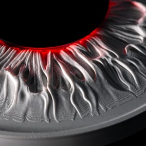 May include: A circular, three-dimensional relief sculpture depicting a human iris with intricate, radiating white ridges and valleys. A deep black pupil sits at the center, surrounded by a vibrant, glowing red ring that separates the pupil from the iris.