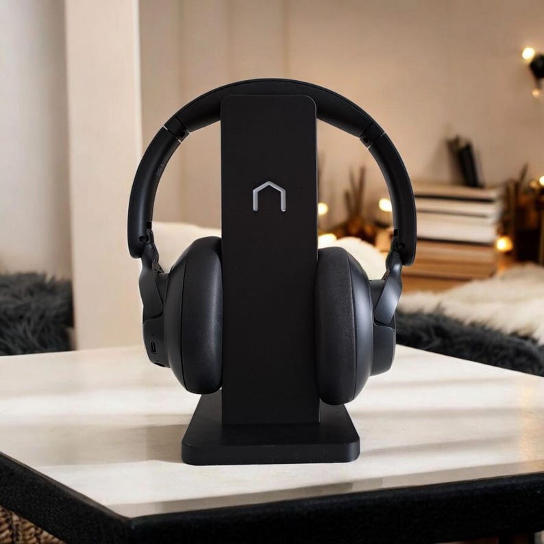 Puede incluir: Auriculares negros sobre la oreja colocados en un soporte negro. El soporte tiene forma rectangular con un peque&ntilde;o logotipo blanco en la parte superior. Los auriculares y el soporte est&aacute;n sobre una superficie blanca.