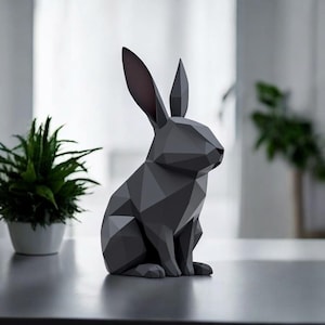 May include: A geometric, dark gray rabbit sculpture with long ears and a red nose. The modern art piece is sitting on a table next to a potted plant. The rabbit is made of many flat, triangular surfaces.
