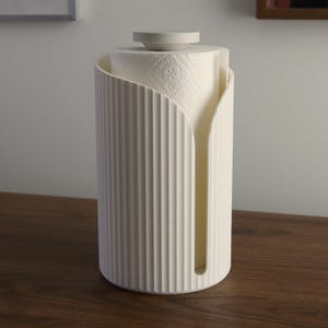 May include: A cream-colored cylindrical paper towel holder featuring a vertical ribbed texture and a wide front opening. The holder has a flat circular top cap and contains a roll of white paper towels with a subtle embossed floral pattern.