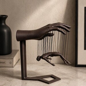 May include: A bronze-colored sculpture depicting a hand holding strings attached to a human figure. The sculpture is mounted on a stand with a geometric base. The artwork evokes themes of control and manipulation.