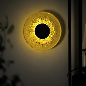 May include: A circular wall lamp featuring a solid black center disc surrounded by a translucent, textured yellow ring that radiates light, creating a sun-like appearance against the wall surface with a warm, glowing illumination effect.