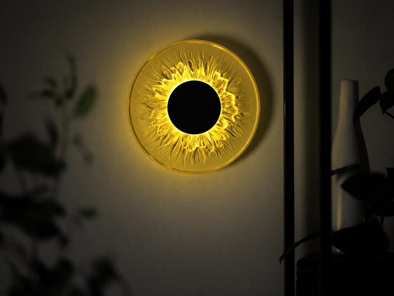 May include: A circular wall lamp featuring a solid black center disc surrounded by a translucent, textured yellow ring that radiates light, creating a sun-like appearance against the wall surface with a warm, glowing illumination effect.
