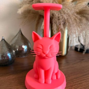 May include: A bright neon pink paper towel holder featuring a sculpted cat figure at the base. The cat has closed eyes and whiskers, sitting upright on a circular platform. A vertical rod extends from the cat's back to a flat top handle.