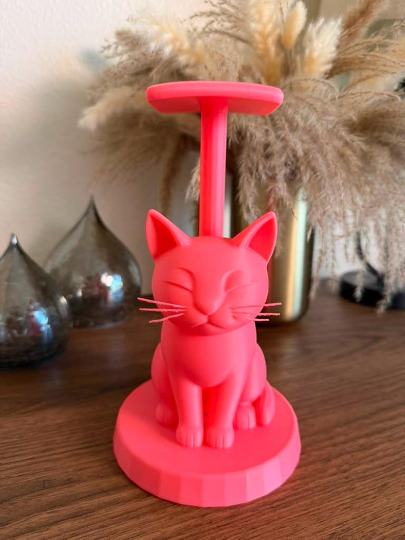 May include: A bright neon pink paper towel holder featuring a sculpted cat figure at the base. The cat has closed eyes and whiskers, sitting upright on a circular platform. A vertical rod extends from the cat's back to a flat top handle.