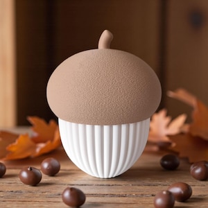 May include: A decorative acorn-shaped object with a textured brown top and a white, ribbed base. The acorn is set on a wooden surface with scattered brown objects and autumn leaves, evoking a fall theme.