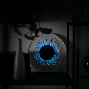 May include: A circular glass art piece featuring a central black disc surrounded by a textured, petal-like pattern illuminated with a vibrant blue light. The object is positioned on a shelf next to a white vase and a dark, spherical glass ornament.