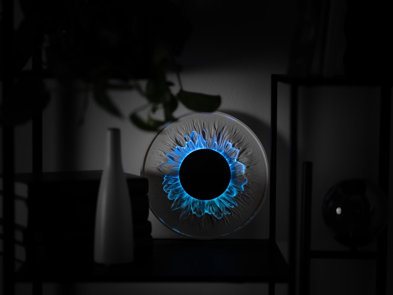 May include: A circular glass art piece featuring a central black disc surrounded by a textured, petal-like pattern illuminated with a vibrant blue light. The object is positioned on a shelf next to a white vase and a dark, spherical glass ornament.