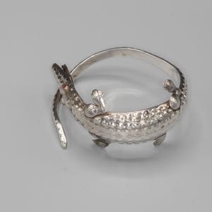May include: A silver-toned bracelet in the shape of an alligator. The alligator's body forms the bracelet's band, with textured scales and detailed legs. The alligator's head features a red gem eye and open jaws.