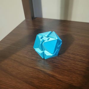 May include: A cyan-colored, geometric puzzle toy with multiple triangular and rectangular facets. The puzzle is resting on a dark brown wooden surface. The toy appears to be 3D-printed.