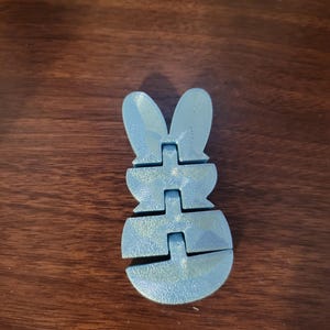 May include: A light blue, articulated bunny figure. The bunny is made of several connected pieces, allowing it to be posed. The figure has a textured surface and is sitting on a dark brown wooden surface. The bunny's ears are upright, and its body is segmented.