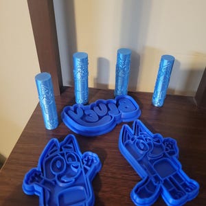 May include: Blue cookie cutters and stamps featuring cartoon characters and the word "Bluey". The set includes character shapes and cylindrical stamps with detailed designs. These tools are likely for baking or crafting.