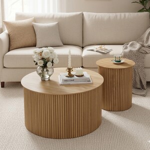 May include: A light-colored, modern living room scene featuring a beige sofa with throw pillows, two wooden coffee tables, and a rug. The round coffee tables have a ribbed design. A vase of white flowers and decorative items are on the tables.