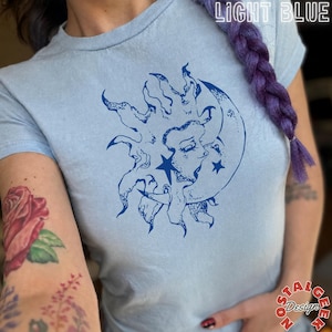 Puede incluir: Una camiseta azul claro con un estampado gráfico azul oscuro de un sol y una luna creciente con rostros y estrellas. El texto "LIGHT BLUE" aparece en la esquina superior derecha y "nostalgic Design" en la esquina inferior derecha.