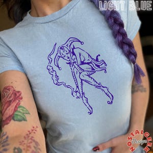 Puede incluir: Una camiseta azul claro con un estampado gráfico de color morado oscuro que muestra una figura de bufón en una pose dinámica. El texto "LIGHT BLUE" aparece arriba a la derecha y "nostal design" abajo a la derecha.