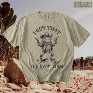 Puede incluir: Una camiseta color caqui con un gráfico marrón oscuro de una rana con sombrero de vaquero y dos pistolas. El texto sobre la rana dice I GOT THAT y el texto debajo dice YEE HAW'TISM. La camiseta tiene cuello redondo y mangas cortas.