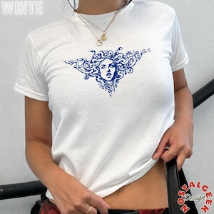 Puede incluir: Una camiseta blanca de manga corta con un estampado gráfico azul de una figura similar a Medusa con serpientes por cabello. La palabra WHITE está impresa en la esquina superior izquierda. La camiseta tiene cuello redondo y una cadena dorada.