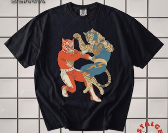 May include: A black short-sleeved t-shirt featuring a graphic of two anthropomorphic tiger wrestlers in masks. One wrestler wears a red and white outfit with stars, while the other wears a blue and gold outfit with a lightning bolt design.