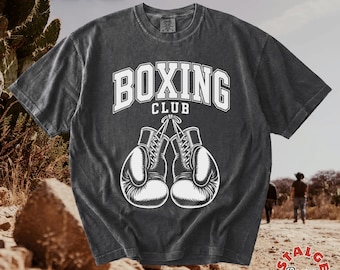 Könnte beinhalten: Ein anthrazitfarbenes Kurzarm-T-Shirt mit einem weißen Grafikdruck von zwei zusammengebundenen Boxhandschuhen unter dem Text BOXING CLUB in einer geschwungenen Schriftart. Das T-Shirt hat einen Rundhalsausschnitt und eine verwaschene Stoffoptik.