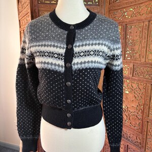 Könnte beinhalten: Eine langärmelige Strickjacke mit einem schwarz-grau-weißen Fair-Isle-Muster auf Brust und Schultern. Das Kleidungsstück hat einen Rundhalsausschnitt, eine durchgehende Knopfleiste mit sechs runden Metallknöpfen sowie schwarze Rippbündchen an Ärmeln und Saum.