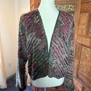 May include: A long-sleeved open-front cardigan featuring a textured, layered design with alternating diagonal stripes of dark green, deep magenta, and multi-colored flecked fabric. The garment has a V-neckline and a solid dark green trim along the edges.