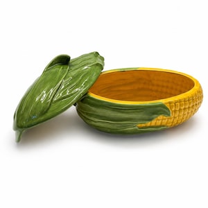 May include: A ceramic serving dish shaped like an ear of corn with a removable lid. The exterior features textured green husks and yellow kernels, while the interior is a smooth, solid yellow. The lid is also shaped like green corn husks.