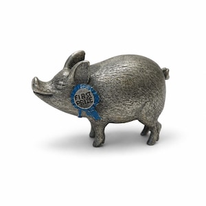 May include: A metallic silver-toned pig figurine with a textured surface, standing in profile. The pig features a blue rosette ribbon attached to its neck, which has the words FIRST PRIZE embossed in black lettering on the center of the badge.