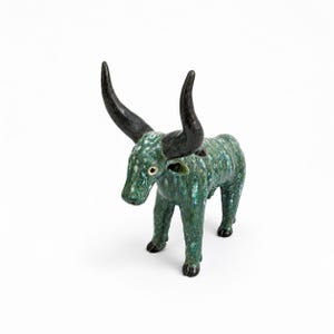 May include: A ceramic bull figurine featuring a mottled green and teal glaze with a glossy finish. The animal has two large, curved, solid black horns, four legs, and a textured surface that mimics a spotted hide, standing in a fixed position.